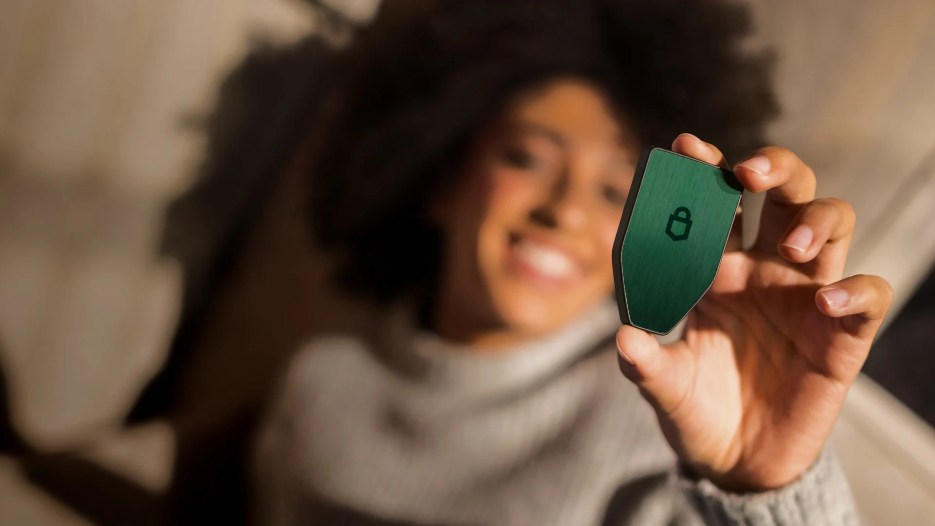 A smiling woman in a cream sweater holds a green Trezor hardware wallet up to the light.
