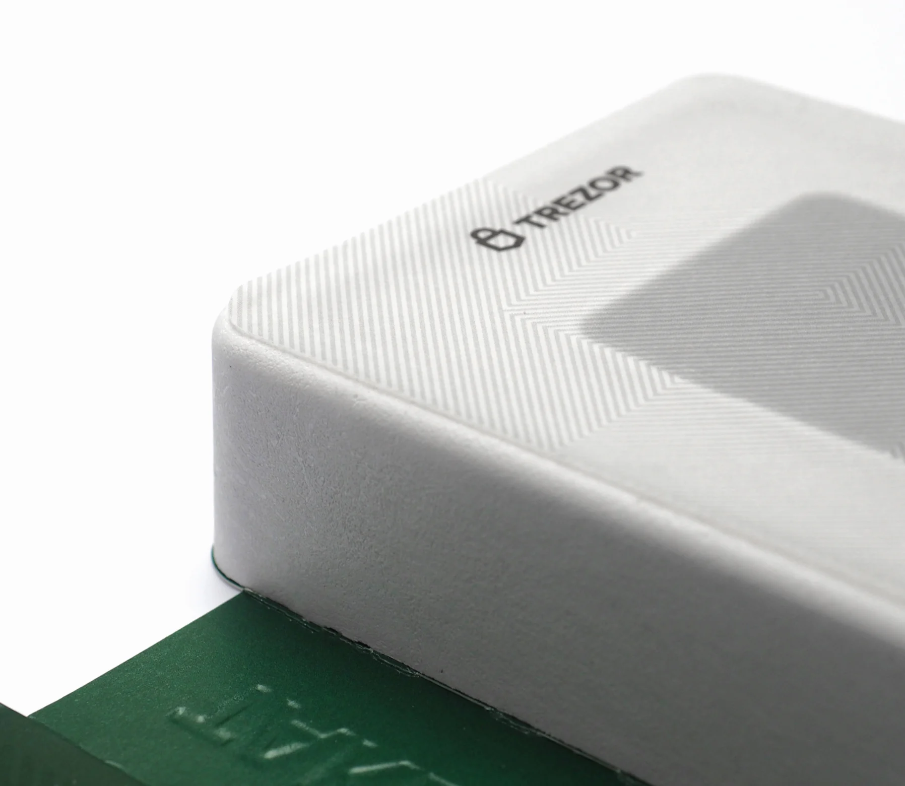 A corner of the white box, its herringbone emboss and Trezor mark catching the light, a green sleeve beneath.